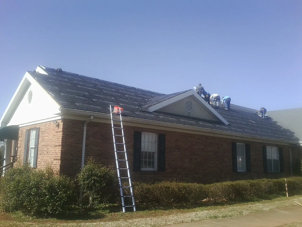 Crew tearing shingles off a brick ranch roof with a ladder set — Metal Roof Repair work by our crew in Barnesville